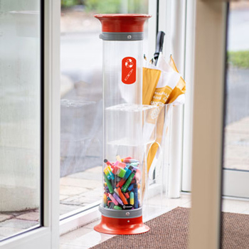 Ein gro&szlig;er, roter, durchsichtiger, rohrartiger Recyclingbeh&auml;lter f&uuml;r E-Zigaretten und Vapes neben einem Fenster